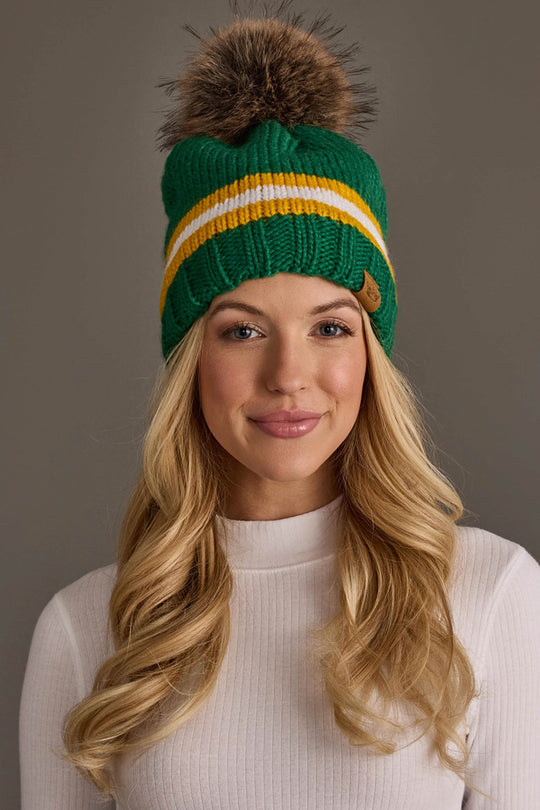 A woman with long blonde hair, smiling in front of a plain gray background, wears the Panache Apparel Collection Green, Gold & White Knit Pom Hat featuring yellow and white stripes and a brown pom-pom paired with a white ribbed top.