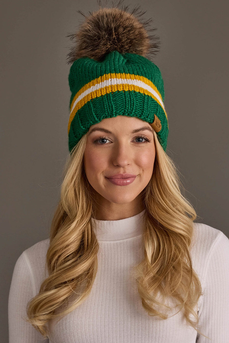 A woman with long blonde hair, smiling in front of a plain gray background, wears the Panache Apparel Collection Green, Gold & White Knit Pom Hat featuring yellow and white stripes and a brown pom-pom paired with a white ribbed top.