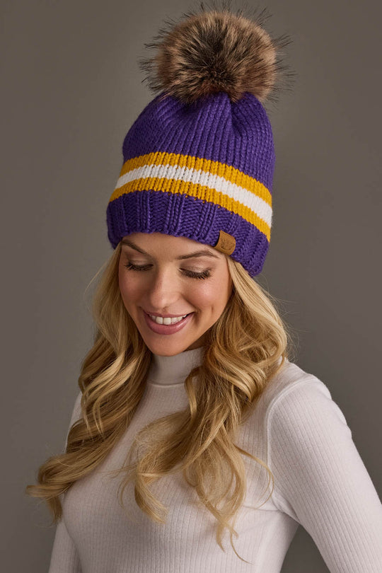 A woman with long blonde hair is smiling and looking down, dressed in a white turtleneck and wearing the Panache Apparel Collection Purple, Gold & White Knit Pom Hat.