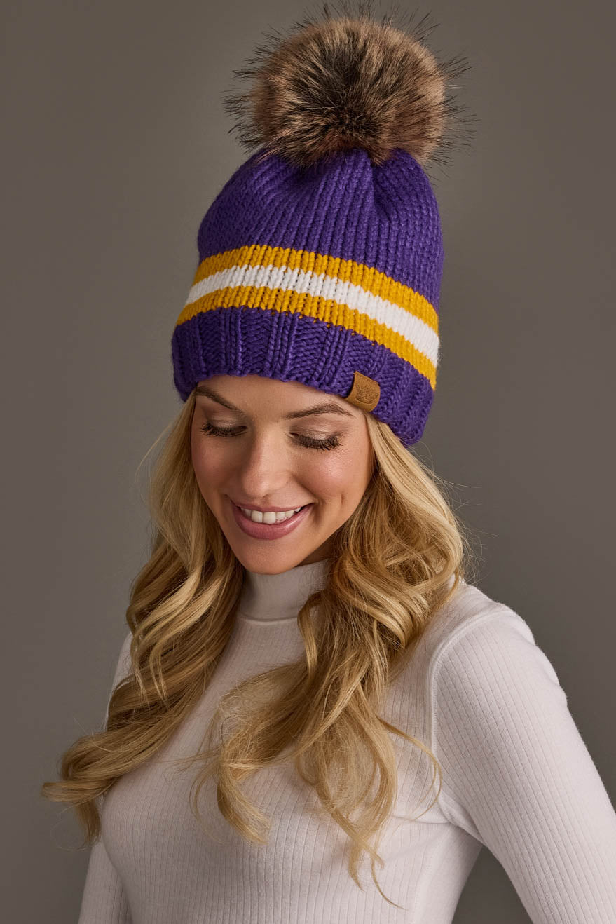 A woman with long blonde hair is smiling and looking down, dressed in a white turtleneck and wearing the Panache Apparel Collection Purple, Gold & White Knit Pom Hat.