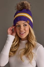 A woman with long blonde hair smiles and touches the Panache Apparel Collection Purple, Gold & White Knit Pom Hat, which features white and yellow stripes with a brown pom-pom, while wearing a white ribbed turtleneck top against a plain gray background.