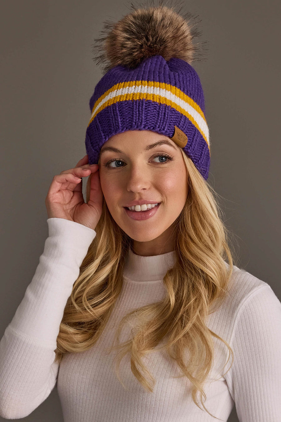 A woman with long blonde hair smiles and touches the Panache Apparel Collection Purple, Gold & White Knit Pom Hat, which features white and yellow stripes with a brown pom-pom, while wearing a white ribbed turtleneck top against a plain gray background.