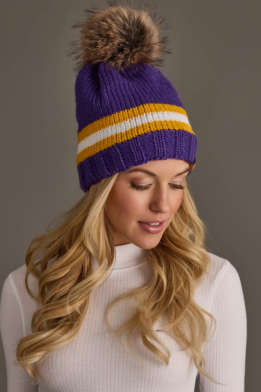A woman with long blonde hair wears the Panache Apparel Collection Purple, Gold & White Knit Pom Hat and a white ribbed turtleneck, looking down and smiling slightly.