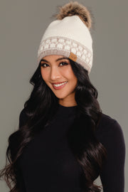 A woman with long black hair, dressed in a black top and smiling, wears the Panache Apparel Collection Ivory & Taupe Patterned Knit Pom Hat, featuring a white knit design with a brown fur pom-pom and patterned band, against a plain gray background.