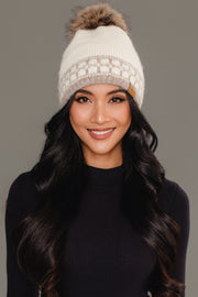 A woman with long black hair is wearing the Panache Apparel Collection’s Ivory & Taupe Patterned Knit Pom Hat and a dark long-sleeve top, smiling in front of a plain gray background.