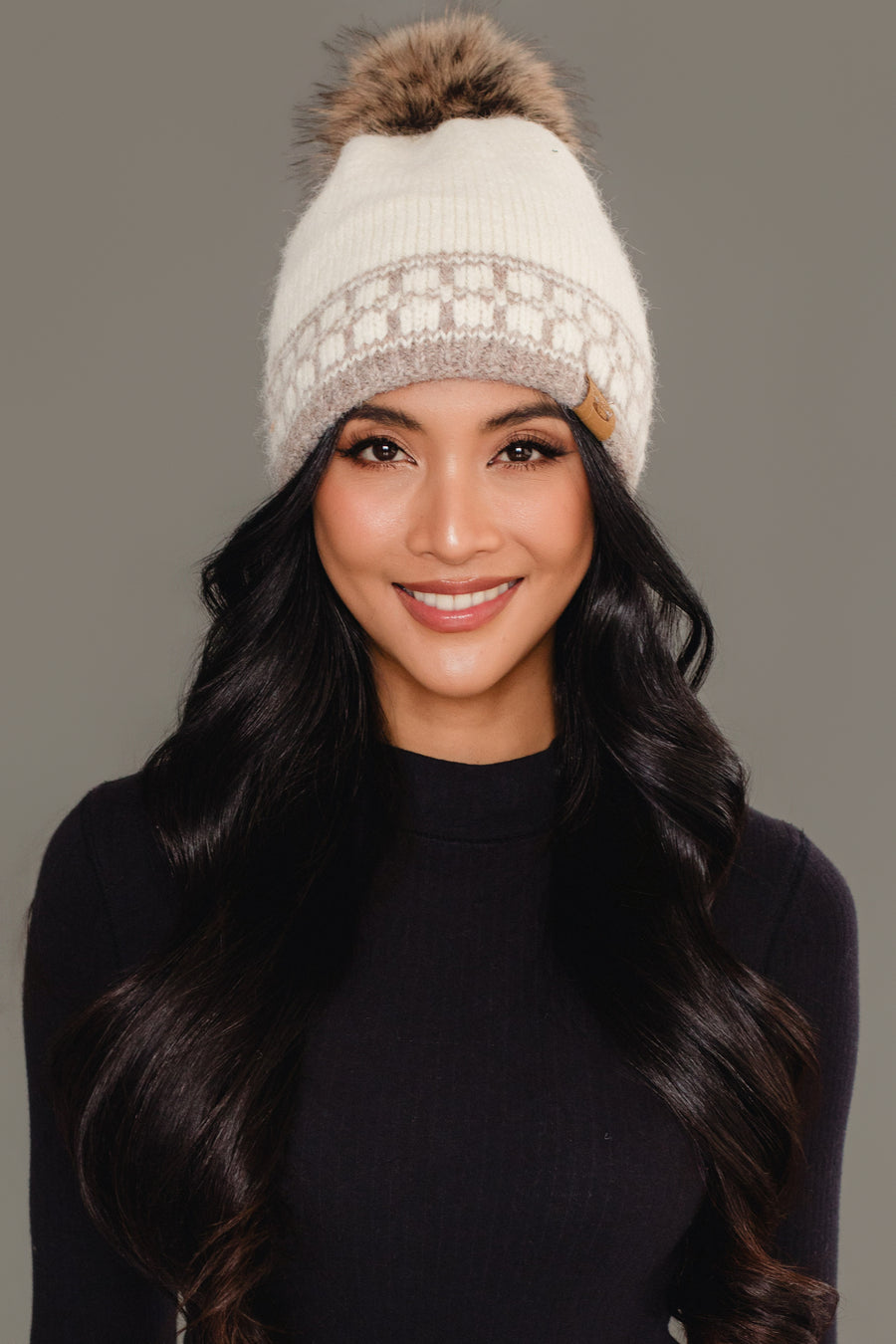 A woman with long black hair is wearing the Panache Apparel Collection’s Ivory & Taupe Patterned Knit Pom Hat and a dark long-sleeve top, smiling in front of a plain gray background.