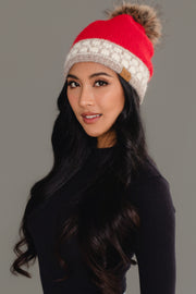 A woman with long, wavy black hair poses against a plain gray background wearing the Panache Apparel Collection’s Red & Taupe Patterned Knit Pom Hat, a red beanie featuring a beige accent band and a fur pom-pom, paired with a black top.