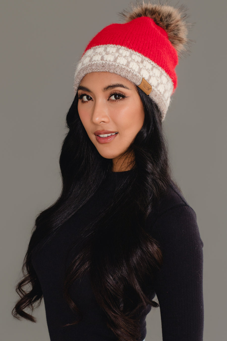 A woman with long, wavy black hair poses against a plain gray background wearing the Panache Apparel Collection’s Red & Taupe Patterned Knit Pom Hat, a red beanie featuring a beige accent band and a fur pom-pom, paired with a black top.