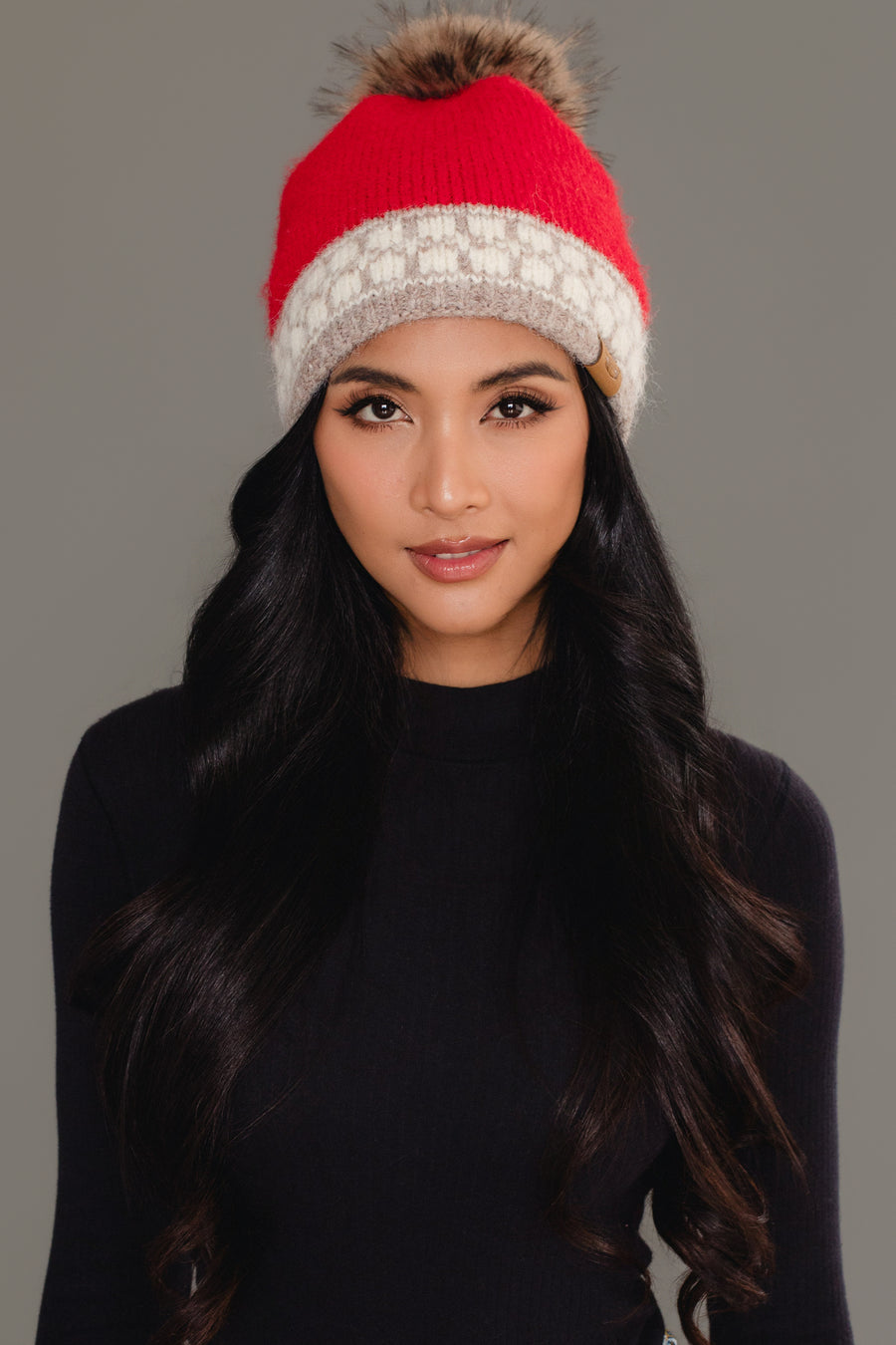 A woman with long dark hair stands against a plain gray background, wearing a black long-sleeve top and the Red & Taupe Patterned Knit Pom Hat by Panache Apparel Collection.