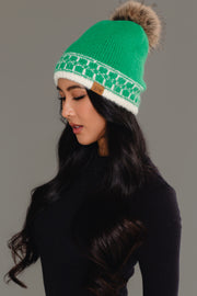 A woman with long dark hair, dressed in a black top, is shown in profile against a neutral background wearing the Panache Apparel Collection’s Green & Cream Patterned Knit Pom Hat, featuring a fur pom-pom.