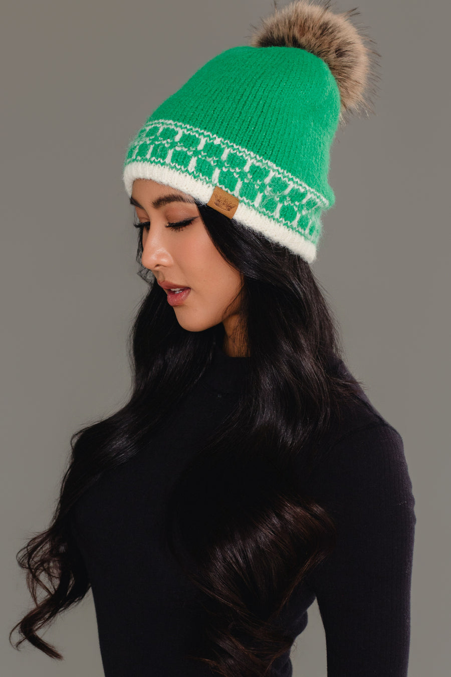 A woman with long dark hair, dressed in a black top, is shown in profile against a neutral background wearing the Panache Apparel Collection’s Green & Cream Patterned Knit Pom Hat, featuring a fur pom-pom.