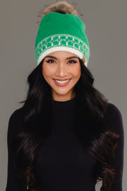 A woman with long dark hair, dressed in a black top, smiles against a plain gray background while wearing the Panache Apparel Collection Green & Cream Patterned Knit Pom Hat, featuring a white pattern and faux fur pom-pom.