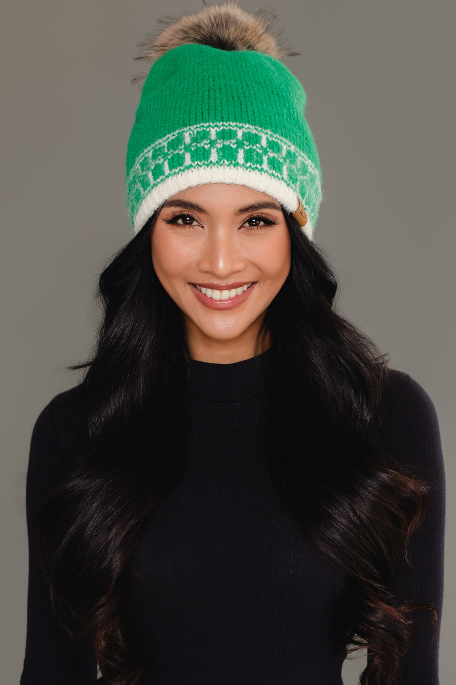 A woman with long dark hair, dressed in a black top, smiles against a plain gray background while wearing the Panache Apparel Collection Green & Cream Patterned Knit Pom Hat, featuring a white pattern and faux fur pom-pom.