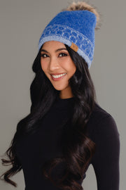 A woman with long, wavy black hair is smiling at the camera against a plain gray background, wearing a black turtleneck and a Panache Apparel Collection Blue & Light Blue Patterned Knit Pom Hat.