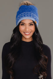 A woman with long black hair smiles at the camera, wearing Panache Apparel Collection’s Blue & Light Blue Patterned Knit Pom Hat, paired with a black top against a plain gray background.