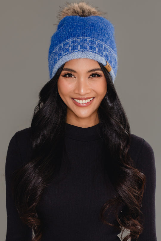 A woman with long black hair smiles at the camera, wearing Panache Apparel Collection’s Blue & Light Blue Patterned Knit Pom Hat, paired with a black top against a plain gray background.