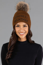 A smiling person wears the Panache Apparel Collection’s Brown & Black Whiskey Weather Pom Hat, featuring a fur pom-pom and embroidered text, paired with a black long-sleeve top against a plain gray background.