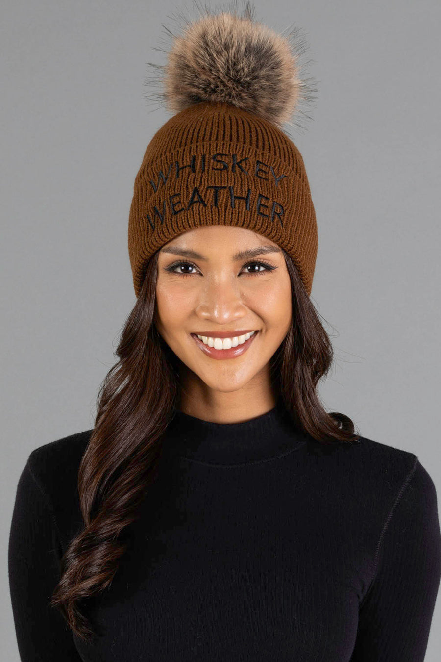 A smiling person wears the Panache Apparel Collection’s Brown & Black Whiskey Weather Pom Hat, featuring a fur pom-pom and embroidered text, paired with a black long-sleeve top against a plain gray background.