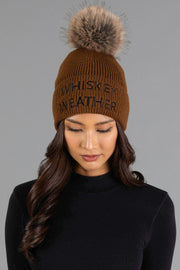 A woman with long, dark wavy hair and a black top is looking down while wearing the Panache Apparel Collection Brown & Black Whiskey Weather Pom Hat, a brown knit beanie featuring a large faux fur pom-pom and “WHISKEY WEATHER” written on the front, against a gray background.
