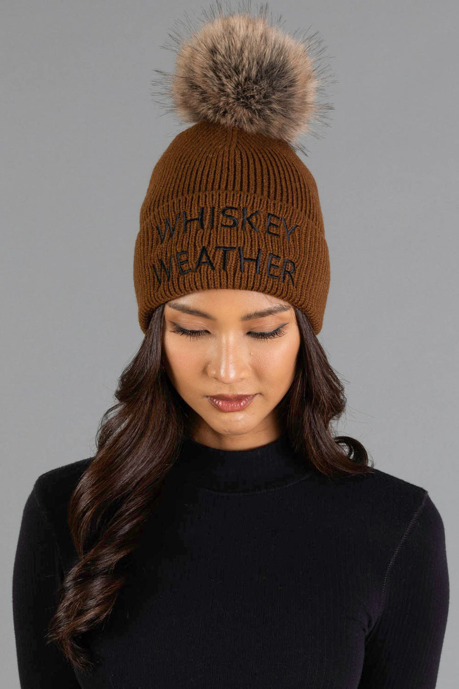 A woman with long, dark wavy hair and a black top is looking down while wearing the Panache Apparel Collection Brown & Black Whiskey Weather Pom Hat, a brown knit beanie featuring a large faux fur pom-pom and “WHISKEY WEATHER” written on the front, against a gray background.