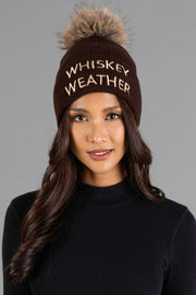 A woman with long wavy hair wears the Panache Apparel Collection Dark Brown & Tan Whiskey Weather Pom Hat, featuring a fur pom-pom and “WHISKEY WEATHER” embroidered on the front, paired with a black sweater against a gray background.