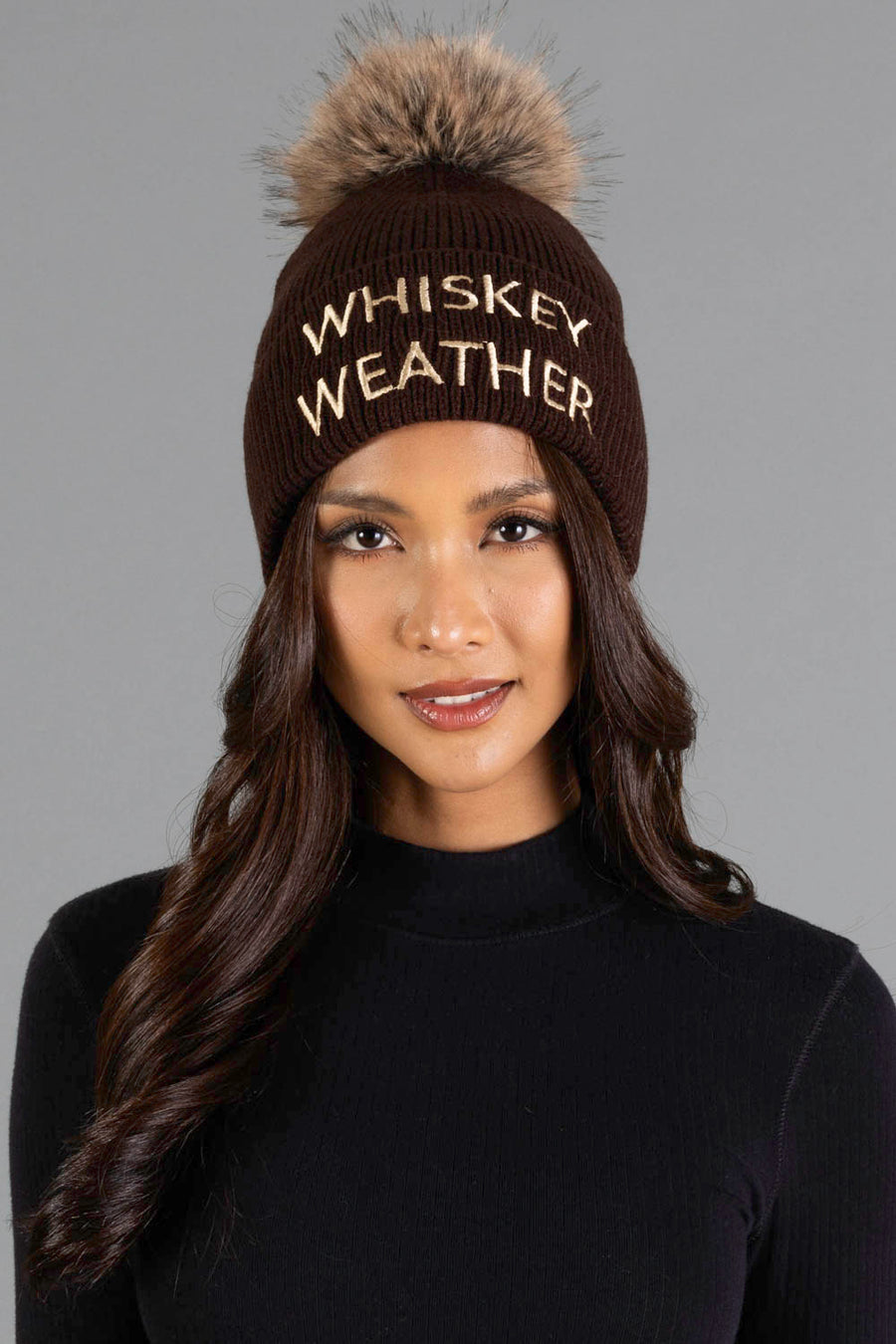 A woman with long wavy hair wears the Panache Apparel Collection Dark Brown & Tan Whiskey Weather Pom Hat, featuring a fur pom-pom and “WHISKEY WEATHER” embroidered on the front, paired with a black sweater against a gray background.