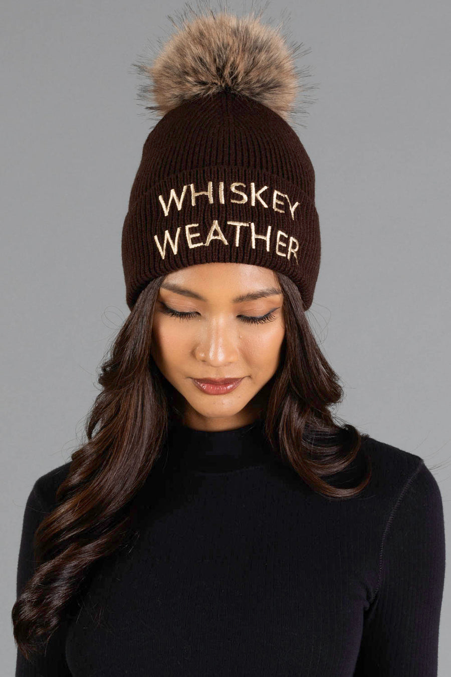 A woman with long brown hair wears the Panache Apparel Collection Dark Brown & Tan Whiskey Weather Pom Hat, a brown knit beanie with a faux fur pom-pom and “WHISKEY WEATHER” on the front, while looking down against a gray background.