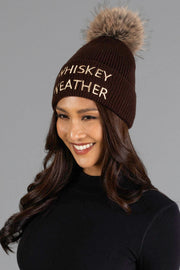A smiling woman wears the Panache Apparel Collection’s Dark Brown & Tan Whiskey Weather Pom Hat, a knit beanie featuring a faux fur pom-pom and “WHISKEY WEATHER” embroidered on the front, paired with a black long-sleeve shirt.