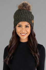 A woman with long, wavy brown hair smiles against a gray background, wearing the Panache Apparel Collection Dark Olive Chenille Pom Hat, a dark knit beanie with a faux fur pom-pom, and a black sweater.