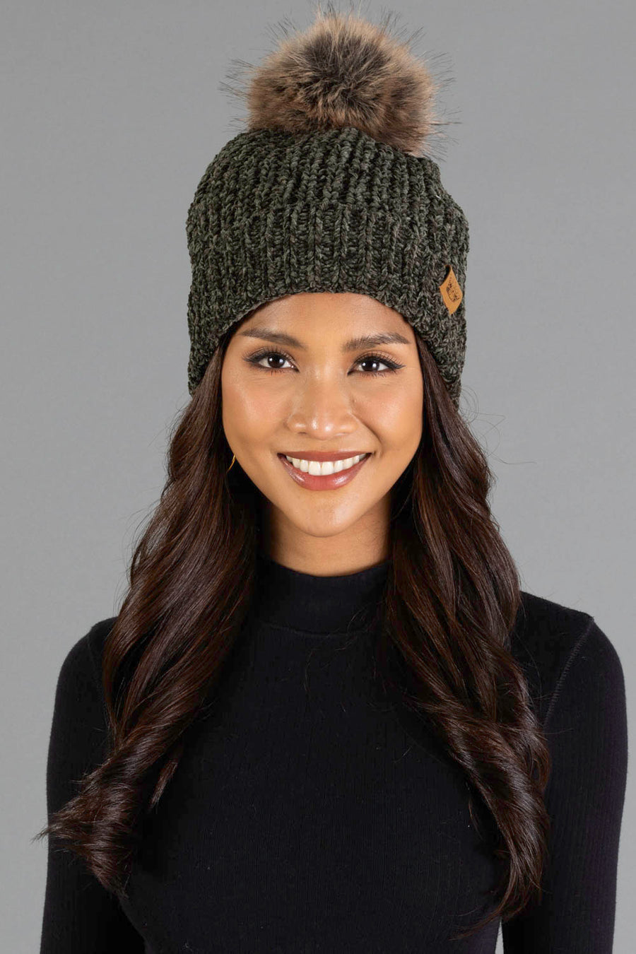 A woman with long, wavy brown hair smiles against a gray background, wearing the Panache Apparel Collection Dark Olive Chenille Pom Hat, a dark knit beanie with a faux fur pom-pom, and a black sweater.