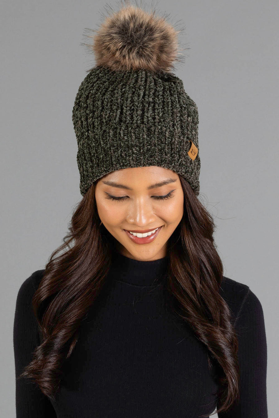 A woman with long wavy hair smiles while wearing the Panache Apparel Collection Dark Olive Chenille Pom Hat, featuring a brown faux fur pom-pom, against a plain gray background.