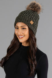 A woman with long wavy hair is smiling and wearing the Panache Apparel Collection Dark Olive Chenille Pom Hat, a textured knit beanie with a furry pom-pom, paired with a black top, set against a plain gray background.