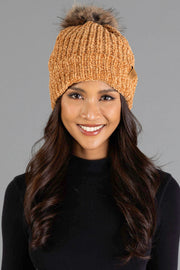 A woman smiles against a plain gray background, dressed in a black sweater and wearing the Panache Apparel Collection Camel Chenille Pom Hat, a knitted orange beanie topped with a fur pom-pom.