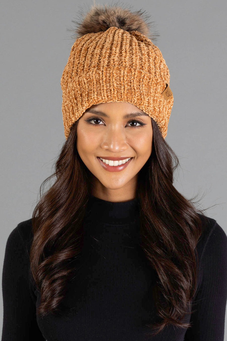 A woman smiles against a plain gray background, dressed in a black sweater and wearing the Panache Apparel Collection Camel Chenille Pom Hat, a knitted orange beanie topped with a fur pom-pom.