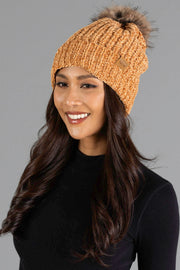 A woman with long wavy dark hair smiles while wearing the Panache Apparel Collection’s Camel Chenille Pom Hat, featuring a faux fur pom-pom, and a black top against a plain gray background.