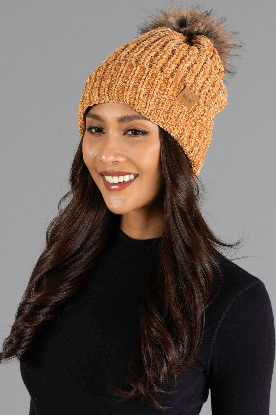 A woman with long wavy dark hair smiles while wearing the Panache Apparel Collection’s Camel Chenille Pom Hat, featuring a faux fur pom-pom, and a black top against a plain gray background.