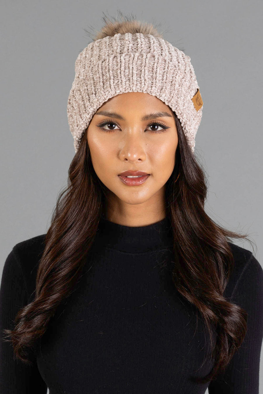 A woman with long dark hair is wearing the Taupe Chenille Pom Hat by Panache Apparel Collection and a black top, posing against a plain gray background.