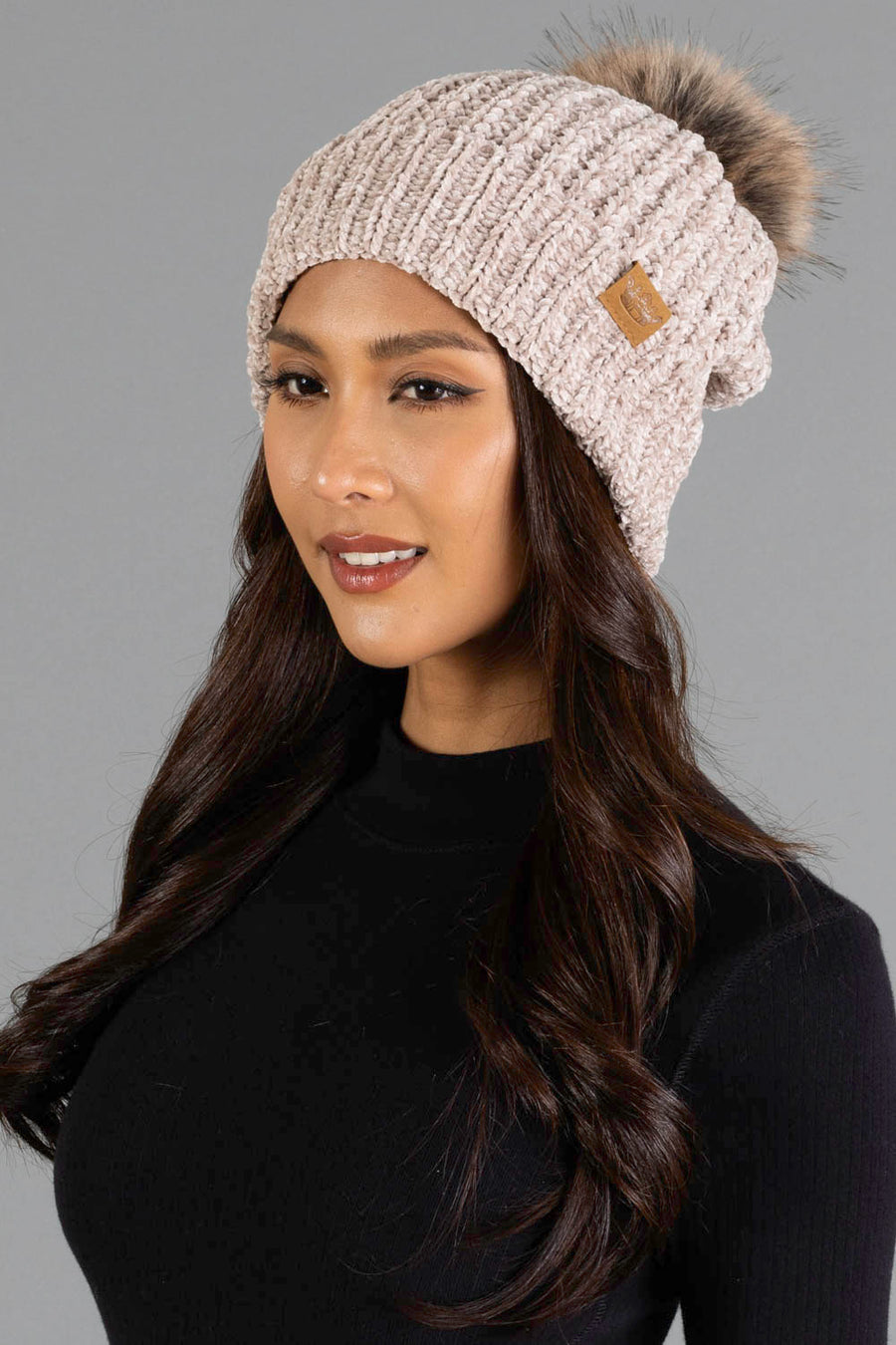 A woman with long dark hair wears the Panache Apparel Collection’s Taupe Chenille Pom Hat, styled with a black top and posing against a plain gray background.