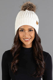 A woman with long dark hair is smiling while wearing the Panache Apparel Collection Ivory Chenille Pom Hat, a white knit beanie with a brown pom-pom, and a black top, posed against a plain gray background.