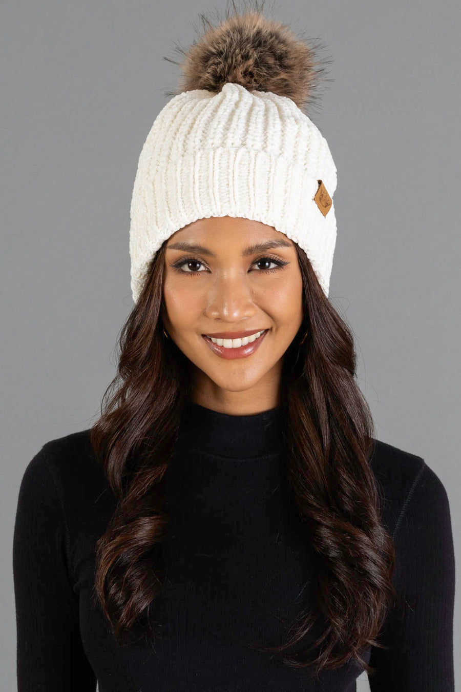 A woman with long dark hair is smiling while wearing the Panache Apparel Collection Ivory Chenille Pom Hat, a white knit beanie with a brown pom-pom, and a black top, posed against a plain gray background.