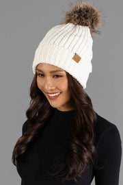 A woman with long dark hair, dressed in a black top and smiling against a gray background, wears the Panache Apparel Collection Ivory Chenille Pom Hat, a white knit beanie featuring a brown faux fur pom-pom.