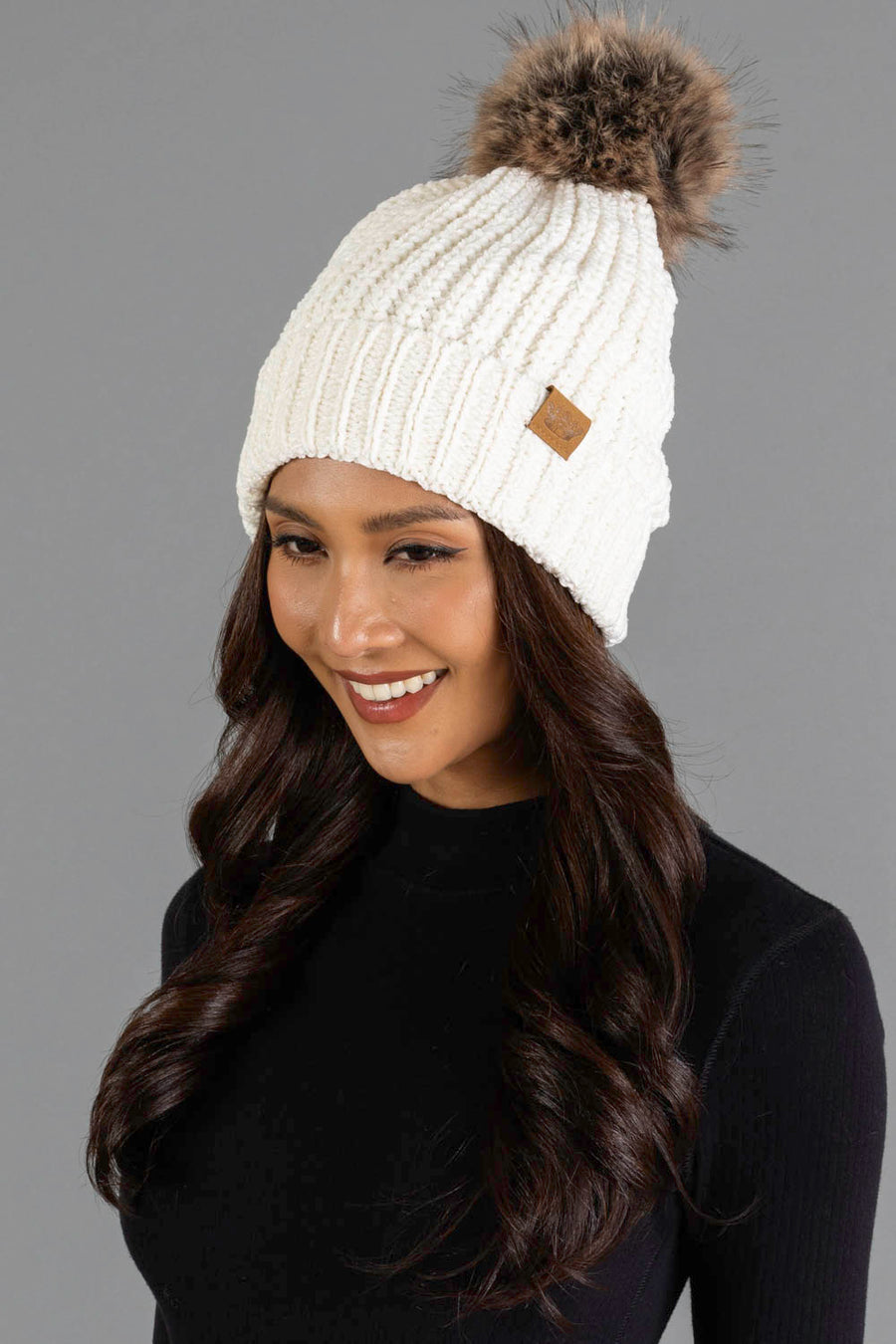 A woman with long dark hair, dressed in a black top and smiling against a gray background, wears the Panache Apparel Collection Ivory Chenille Pom Hat, a white knit beanie featuring a brown faux fur pom-pom.