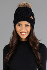A woman with long brown hair wearing the Black Chenille Pom Hat from Panache Apparel Collection and a black sweater smiles at the camera against a gray background.