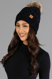 A woman with long dark hair smiles against a plain gray background, wearing the Panache Apparel Collection Black Chenille Pom Hat, a black knit beanie topped with a fur pom-pom, paired with a black turtleneck sweater.