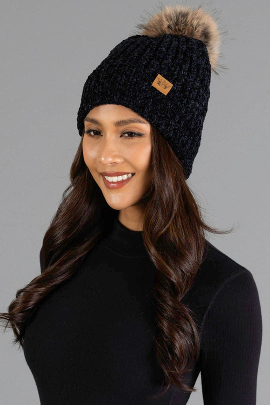 A woman with long dark hair smiles against a plain gray background, wearing the Panache Apparel Collection Black Chenille Pom Hat, a black knit beanie topped with a fur pom-pom, paired with a black turtleneck sweater.
