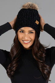 A smiling person with long, wavy hair is wearing the Black Chenille Pom Hat from Panache Apparel Collection, a black knit beanie featuring a faux fur pom-pom, paired with a black long-sleeve top, hands touching the sides of the hat, set against a plain gray background.