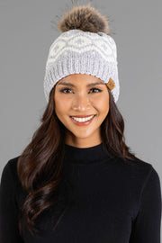 A woman with long dark hair and a black top smiles while wearing the Panache Apparel Collection’s Gray & Cream Patterned Chenille Pom Hat, featuring a plush fur pom-pom, against a plain gray background.