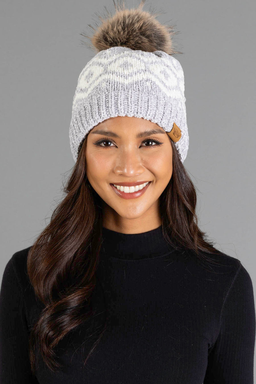 A woman with long dark hair and a black top smiles while wearing the Panache Apparel Collection’s Gray & Cream Patterned Chenille Pom Hat, featuring a plush fur pom-pom, against a plain gray background.