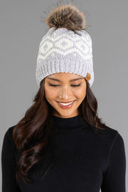 A woman with long dark hair smiles and looks down while wearing the Panache Apparel Collection’s Gray & Cream Patterned Chenille Pom Hat, featuring a white pattern and a large faux fur pom-pom, paired with a black top against a gray background.