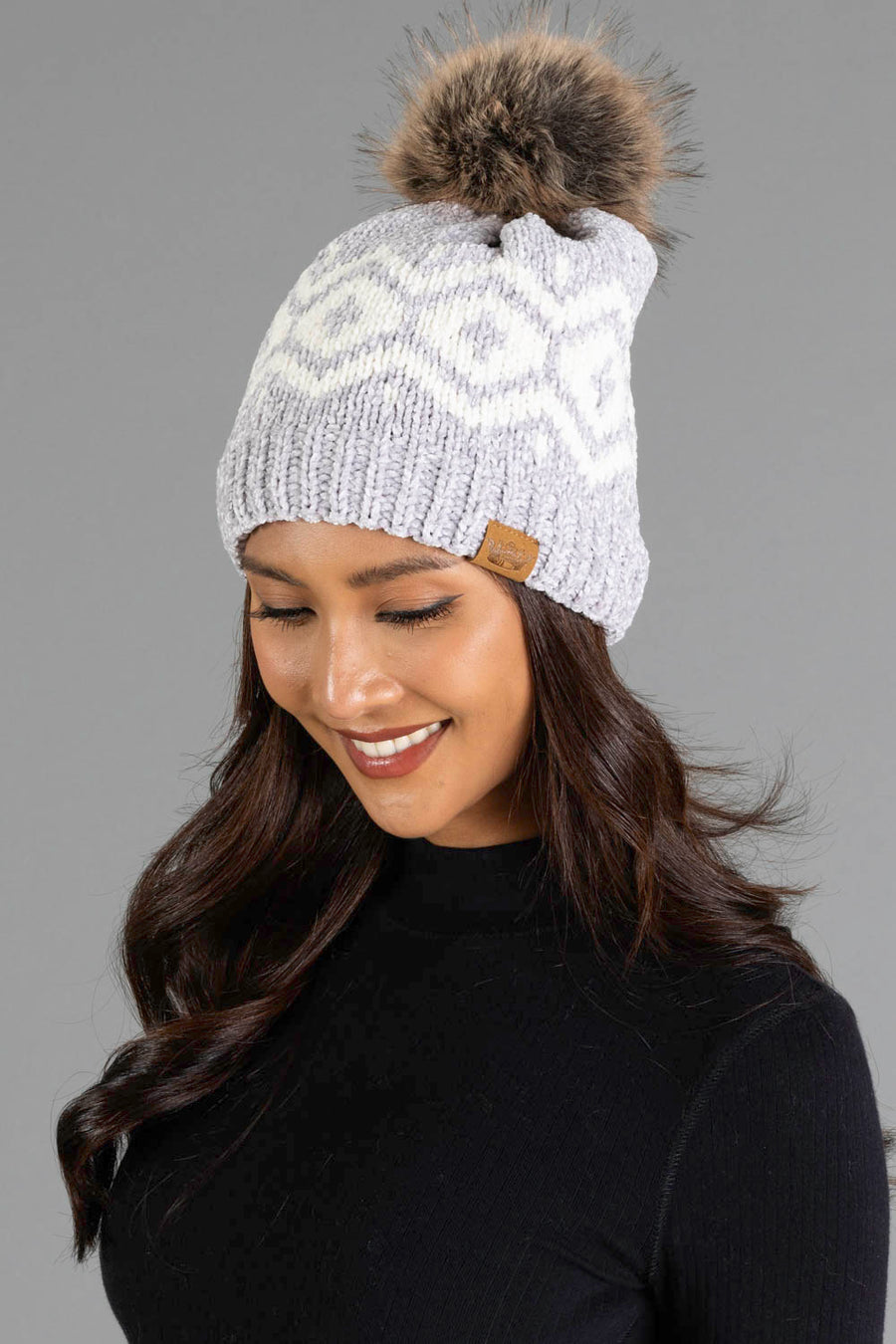 A woman with long brown hair is smiling and looking down while wearing the Panache Apparel Collection Gray & Cream Patterned Chenille Pom Hat, featuring a white zigzag pattern and brown tag. She is dressed in a black top against a gray background.